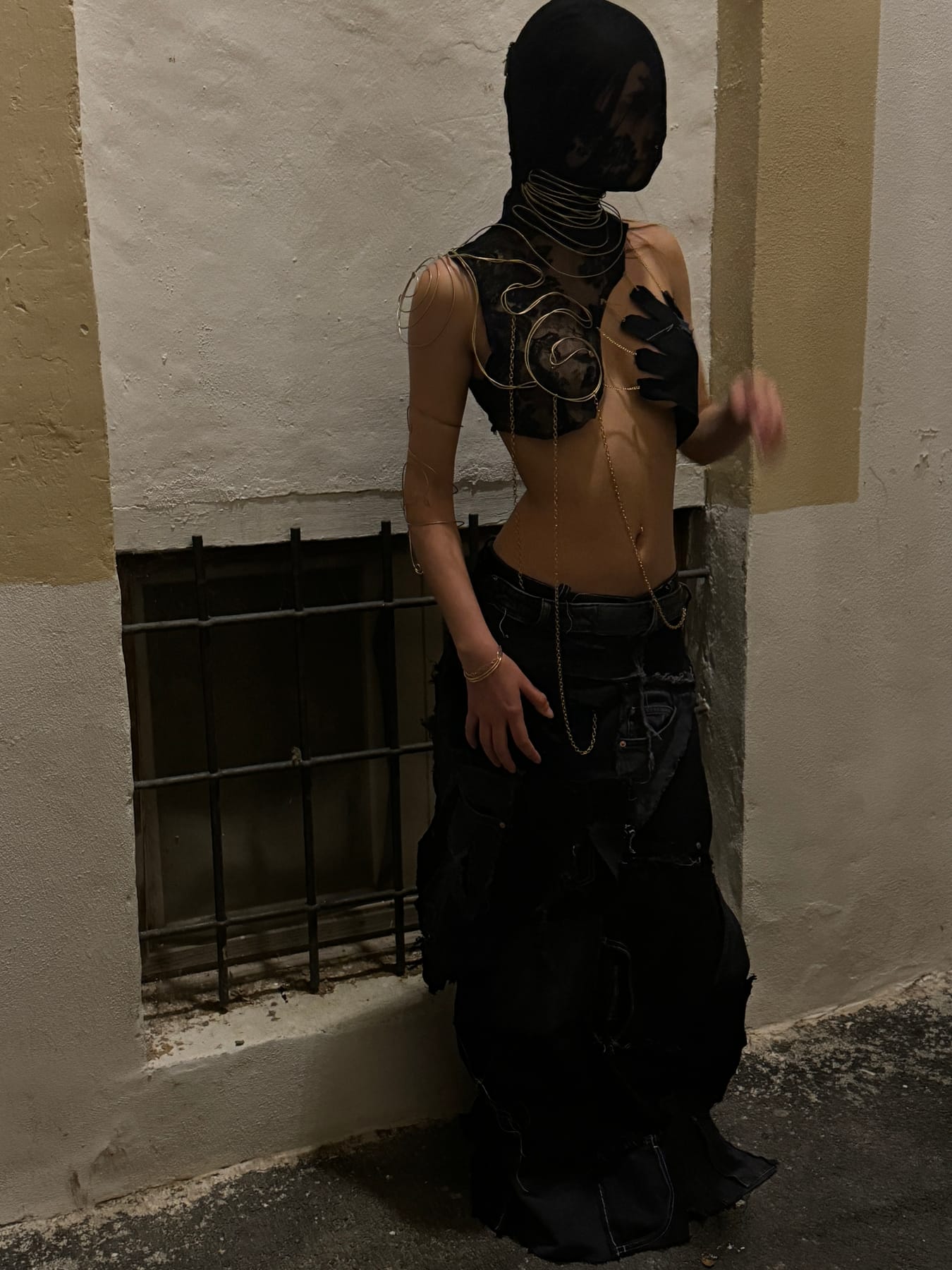 Figura con balaclava de encaje y estructura de alambre dorado en un callejón nocturno